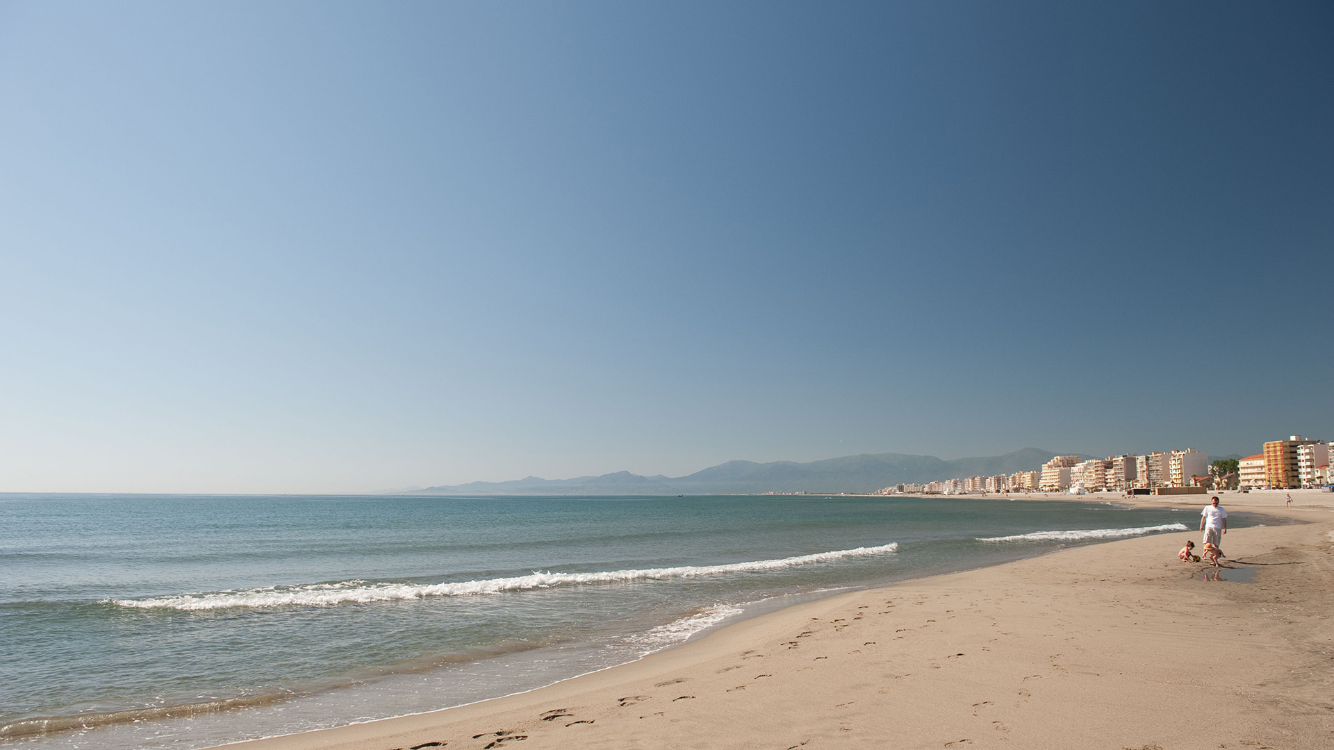 Vue de la plage de Canet-en-Roussillon avec son sable fin, la mer Méditerranée turquoise et la chaîne des Pyrénées au loin.