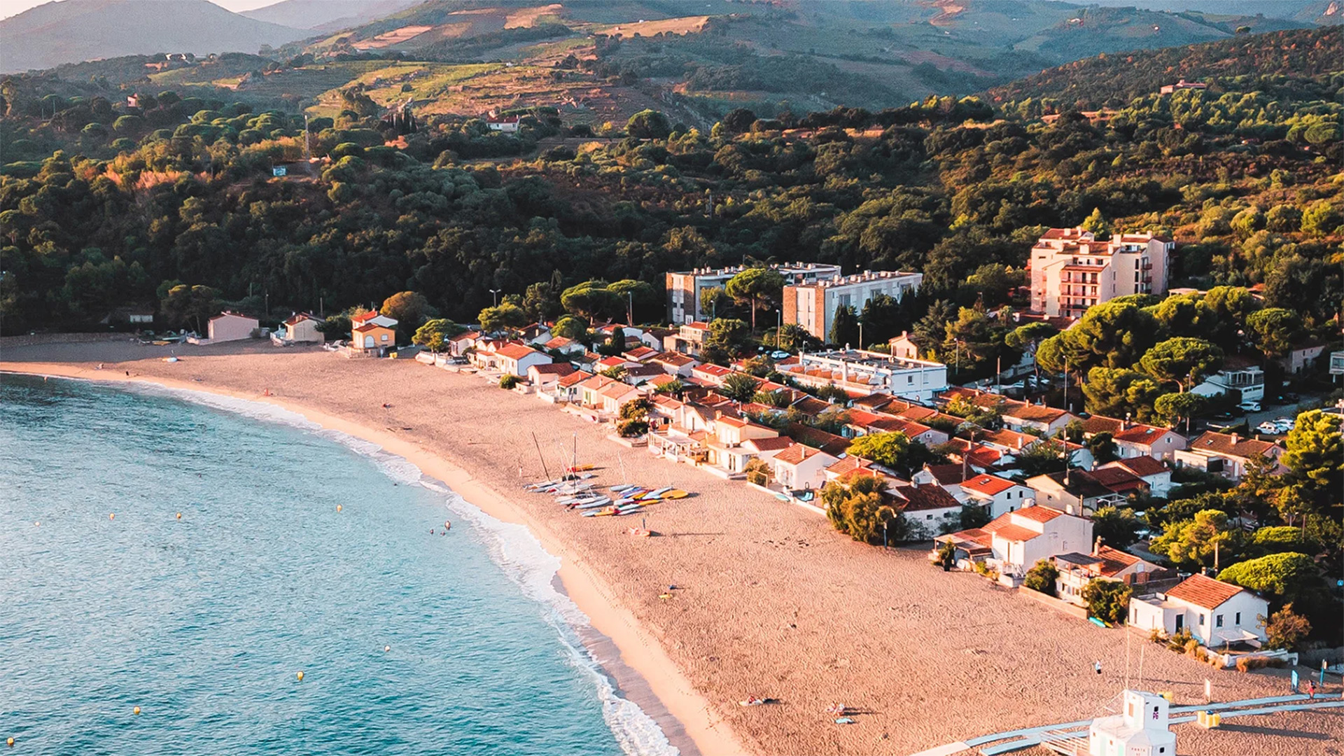 Vue de la plage du Racou à Argelès-sur-Mer avec son sable, ses petites maisons en bord de mer et les montagnes de la Côte Vermeille en arrière-plan.