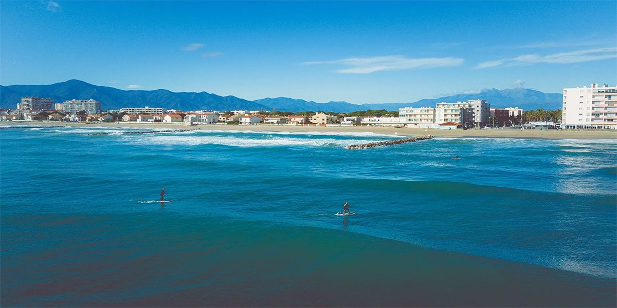 Pratiquants de paddle au large de la plage de Saint-Cyprien avec la Méditerranée, le littoral et le massif des Albères en arrière-plan.